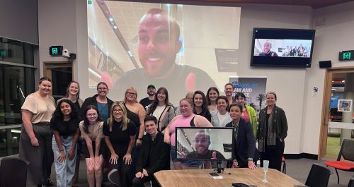 Group of students smiling in front of a computer screen. The computer screen shows a male panel member smiling.