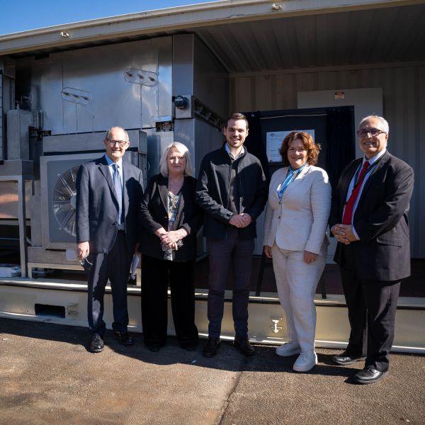 Professor Behdad with a group of people smiling to camera andstanding in front of the metal Hydro Harvester equipment which is the size of a shipping container 