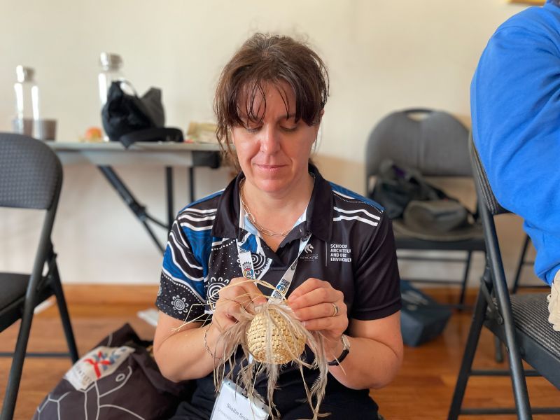 PhD student Shellie Smith sitting on the floor weaving a circle with feathers and straw.
