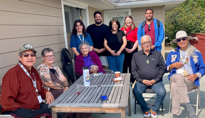 Group of ten people gathered around a wooden table on a concrete porch. Five Elders are sitting in chairs with five delegates standing in the background.