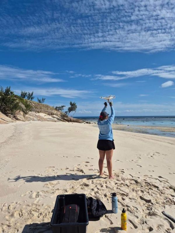 Kristen holding a drone overhead on a sunny beach with clear blue skies and a gentle coastline.