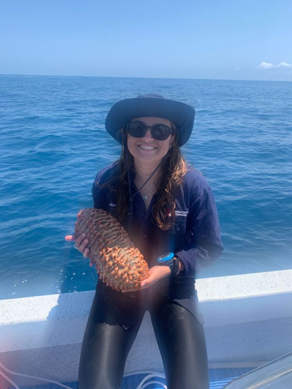 Kristen wears a dark hat and sunglasses, holding a large sea cucumber while sitting on the edge of a boat out at sea.