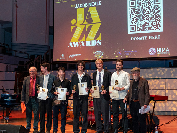 Seven people stand on stage holding awards at the Jacob Neale Jazz Awards, with a large screen behind them showing event details and a QR code for donations.