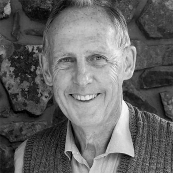 Bob Brown web A grayscale image of Bob Brown wearing a collared shirt and sweater vest, standing in front of a stone wall.