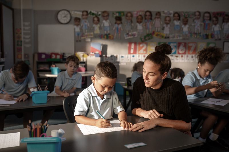 Young boy in a classroom, his teacher sitting next to him helping with his schoolwork