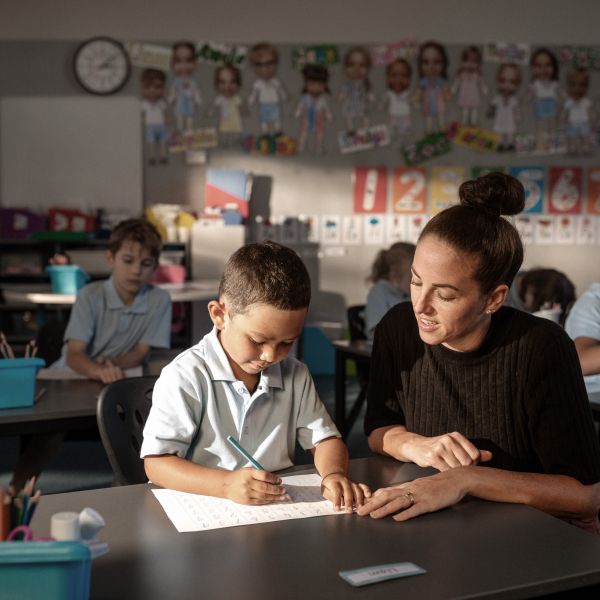 Young boy in a classroom, his teacher sitting next to him helping with his schoolwork