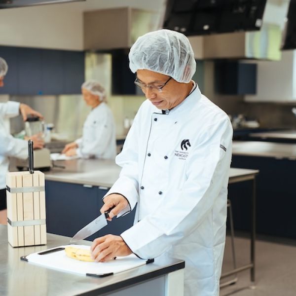 Chef in a white coat and hairnet slices bananas on a cutting board in a professional kitchen. Other chefs work in the background, creating a focused atmosphere.