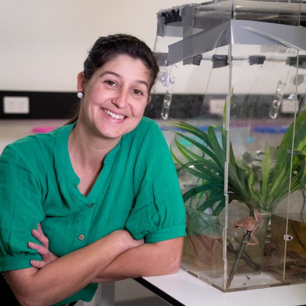 Dr Kaya Klop-Toker pictured in a lab next to a Littlejohn's tree frog behind glass