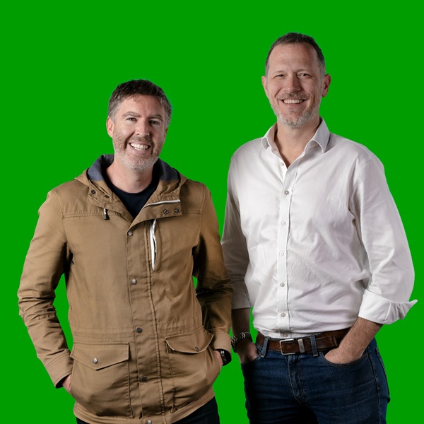 Two men with their hands in their pockets, smiling for the camera in front of a green background