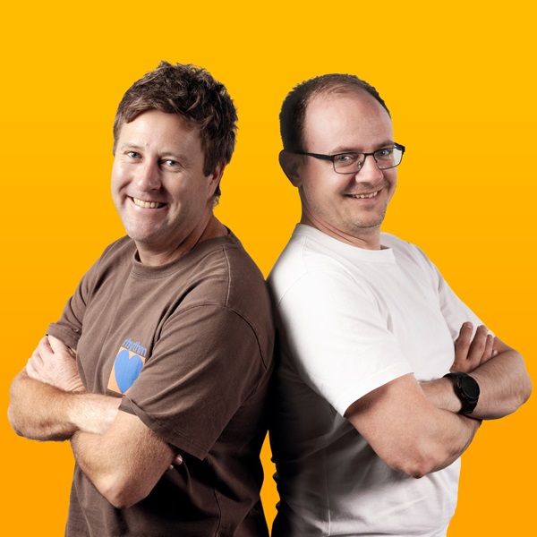 Two men with arms corssed, back-toback, smiling for the camera in front of a yellow background