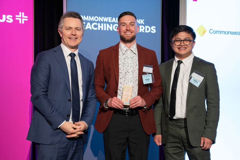 Josh is posing with two other men while holding an award at the Commonwealth Bank Teaching Awards event. They are all smiling and standing in front of a promotional banner.