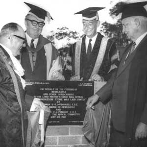 Four men in University gowns and caps standing around a stone marking the opening of the Great Hall