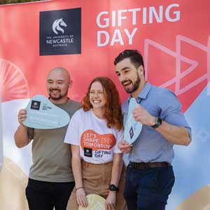 Three people stand in front of a Gifting Day photo wall, smiling and holding placards about Gifting Day. The person in the middle is also wearing a Gifting Day shirt.