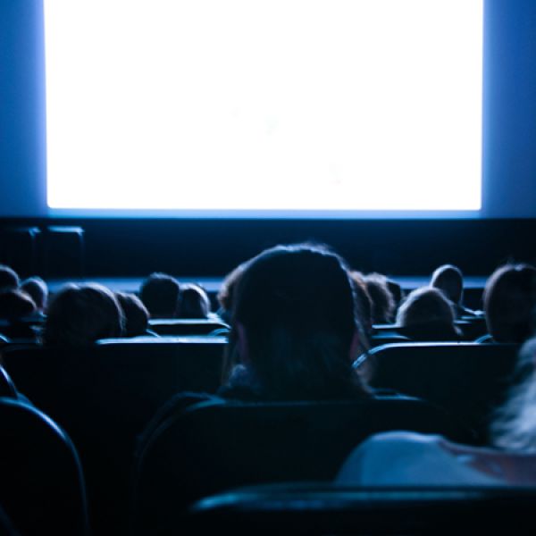 A dimly lit audience sits facing a large, bright movie screen in a theater.
