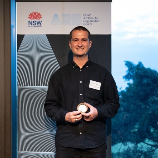 A person smiling while holding an award in front of a NSW Architects Registration Board backdrop. The mood is celebratory and professional.