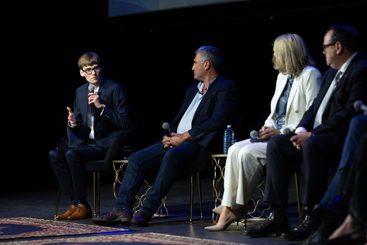 Myles Egan holds a handheld microphone and speaks to panellists, all sitting on chairs on a stage