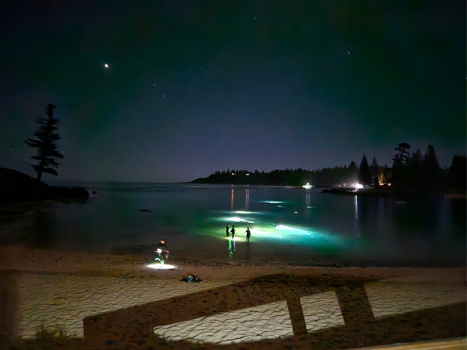 Three people can be seen standing in the ocean at night time, lights illuminate the water from below - coloured blue and green.