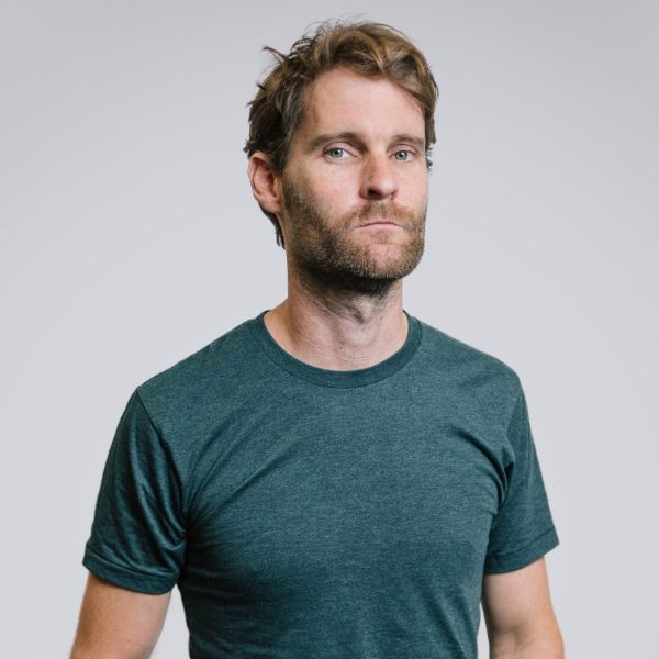 Studio portrait of a person with short, slightly tousled hair and light facial hair, wearing a green T-shirt against a plain gray background.