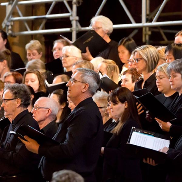 A large choir dressed in black stands in rows holding sheet music folders and singing together on a staged indoor performance setting.
