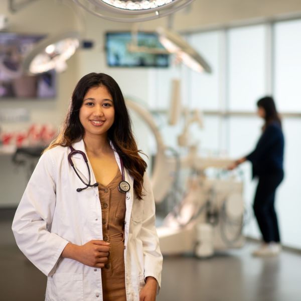 A young woman with brown hair is smiling as she wears a white lab coat and stethoscope around her neck. She is in a clinical medicine setting.