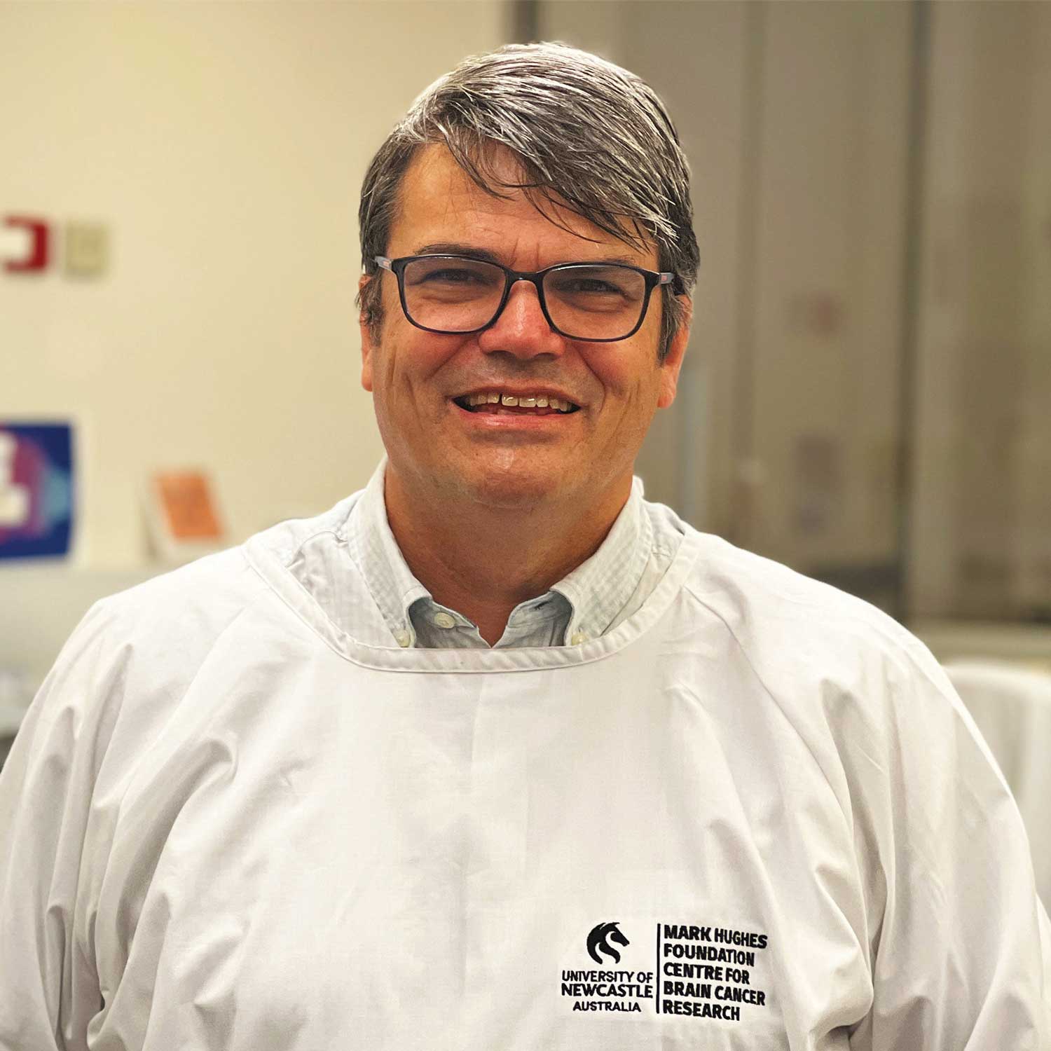 Head-and-shoulders portrait of a person wearing glasses and a white laboratory gown in a research facility. The gown features the University of Newcastle Australia and Mark Hughes Foundation Centre for Brain Cancer Research logos, with laboratory equipment softly visible in the background.
