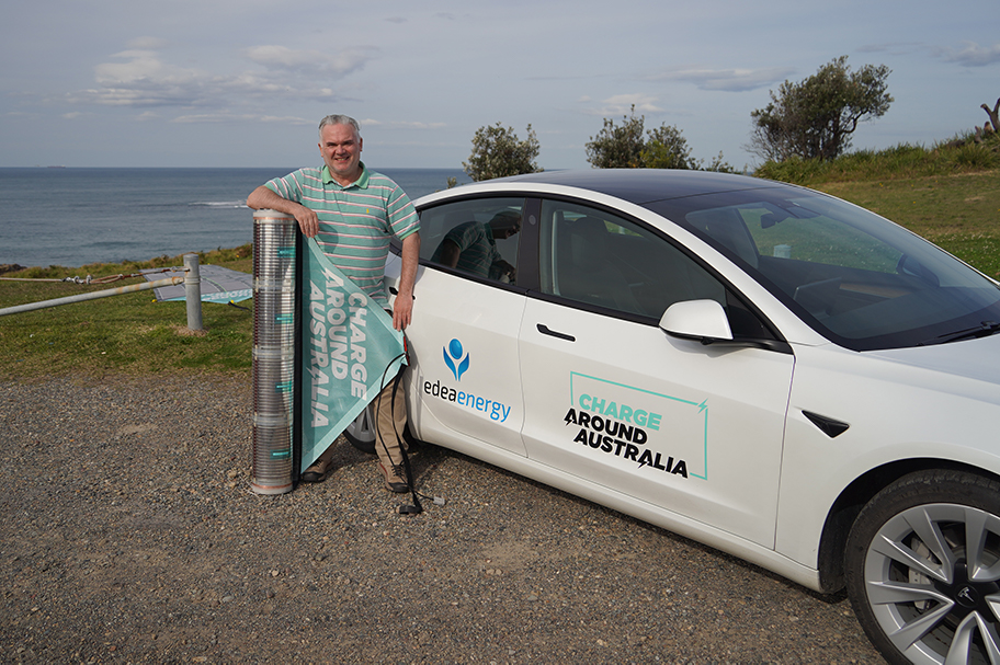Stuart McBain stands next to his car with a roll of printed solar and the beach in the background