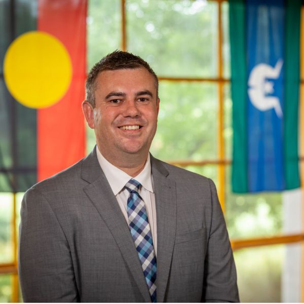 Nathan Towney looks at the camera. The Aboriginal flag and the Torres Strait Islander Flag hangs behind him