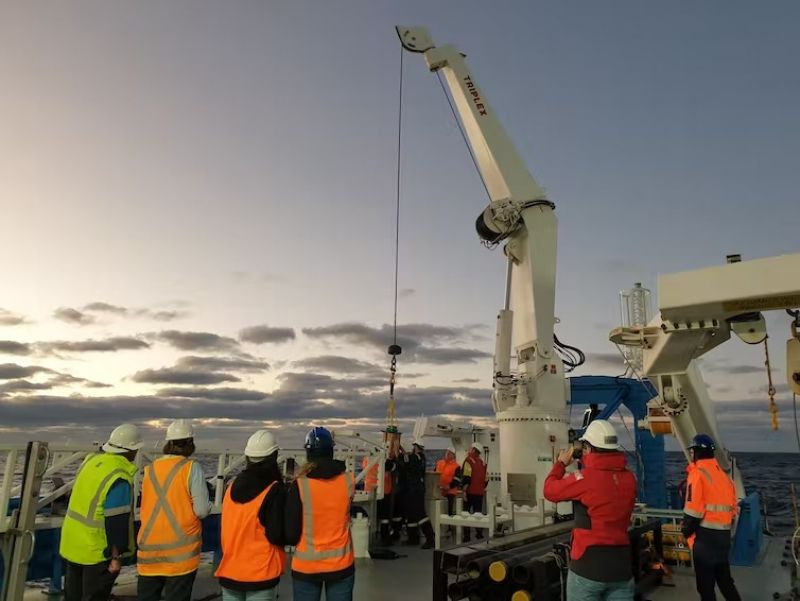A group of researchers with high-vis clothing and hard hats gather around a crane on a boat deck. 
