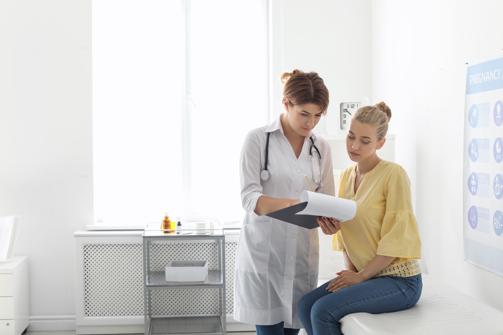 A doctor seeing a patient sitting on a treatment bed in a doctors office with a stethoscope around her neck.