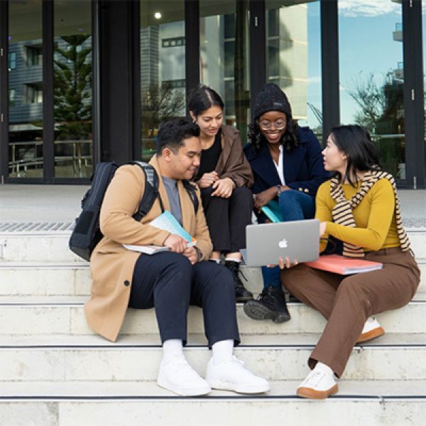 OF Parents and Carers Info Session Four students sitting on the stairs at the entrance of a university building. They are talking to each other and looking at a laptop screen together.