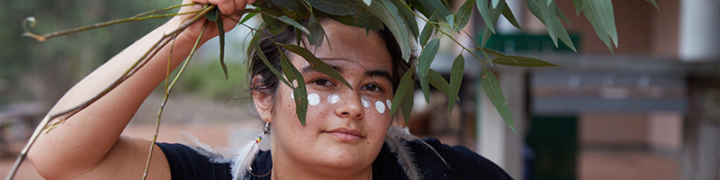 An Aboriginal person dances with some eucalyptus leaves, they have white dots painted across their cheeks