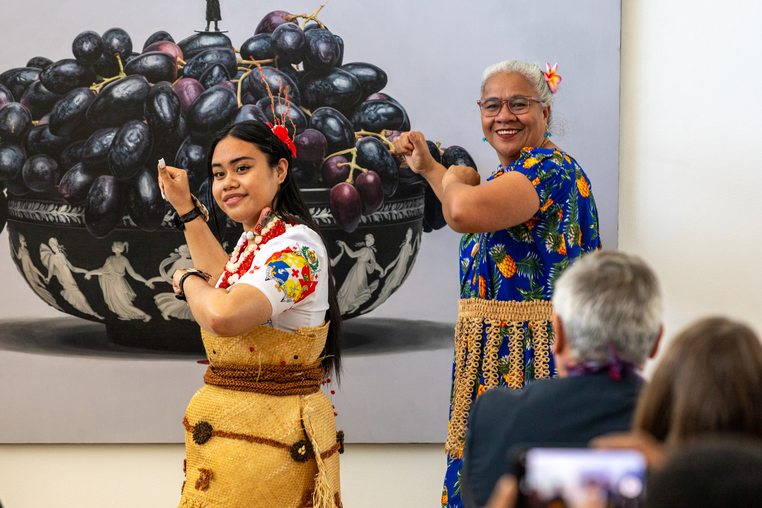 Two women dance wearing traditional Pacific dress in front of a bowl of purple grapes