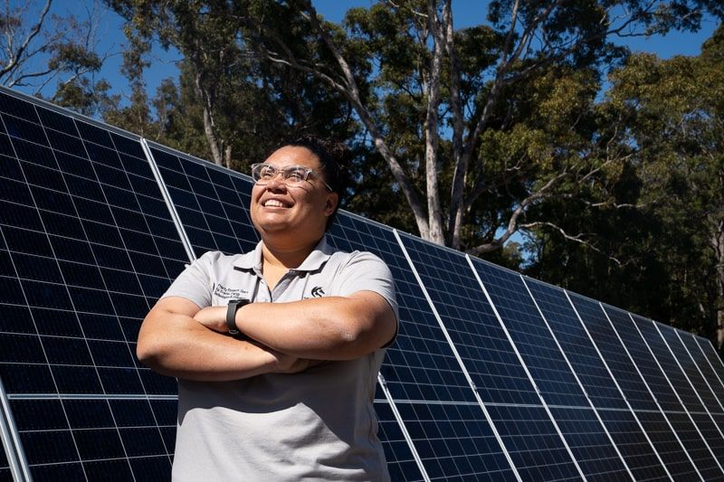 Priscilla stands next to tall solar panels. She looks up at the sun
