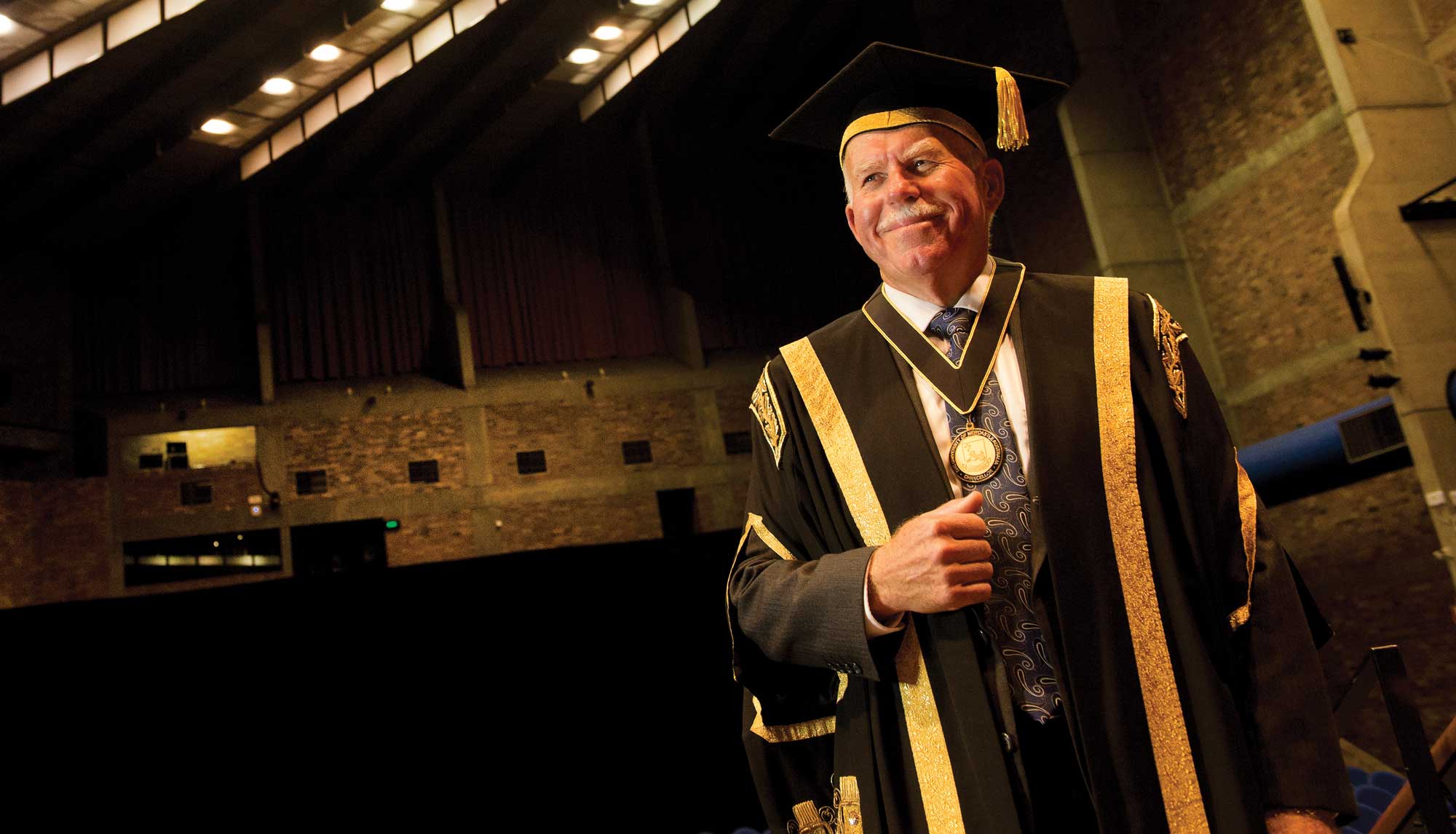 Man in ceremonial graduation robes, smiling in a large and dark brick building