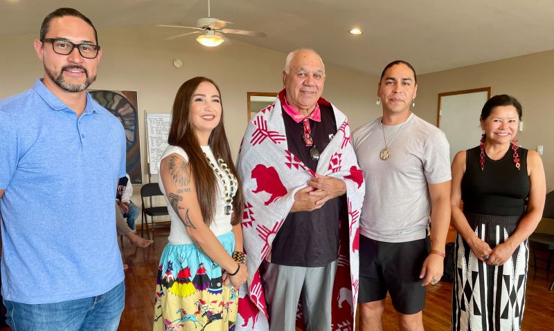 Photo of five people standing next to each other with Professor Bob Morgan standing in the middle with a white blanket with a red pattern of symbols and bison around his shoulders.