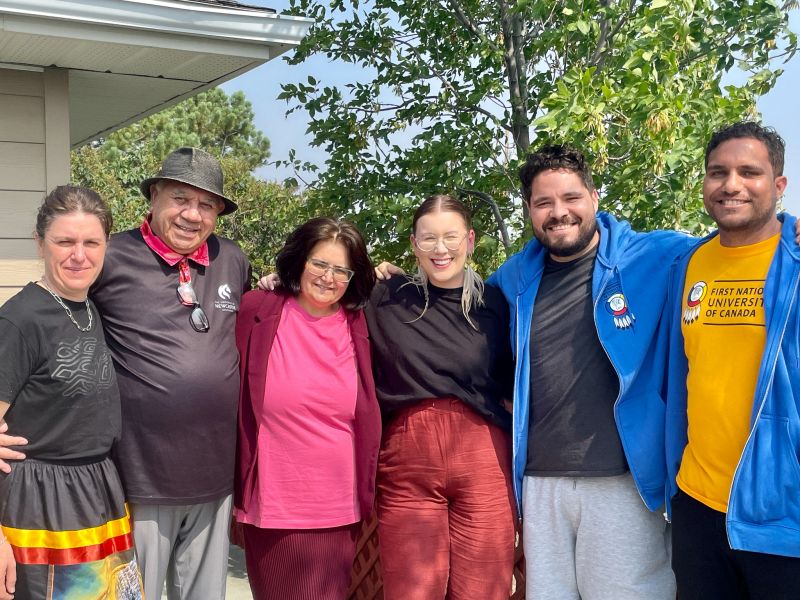 Group of five people standing in a row with trees in the background.