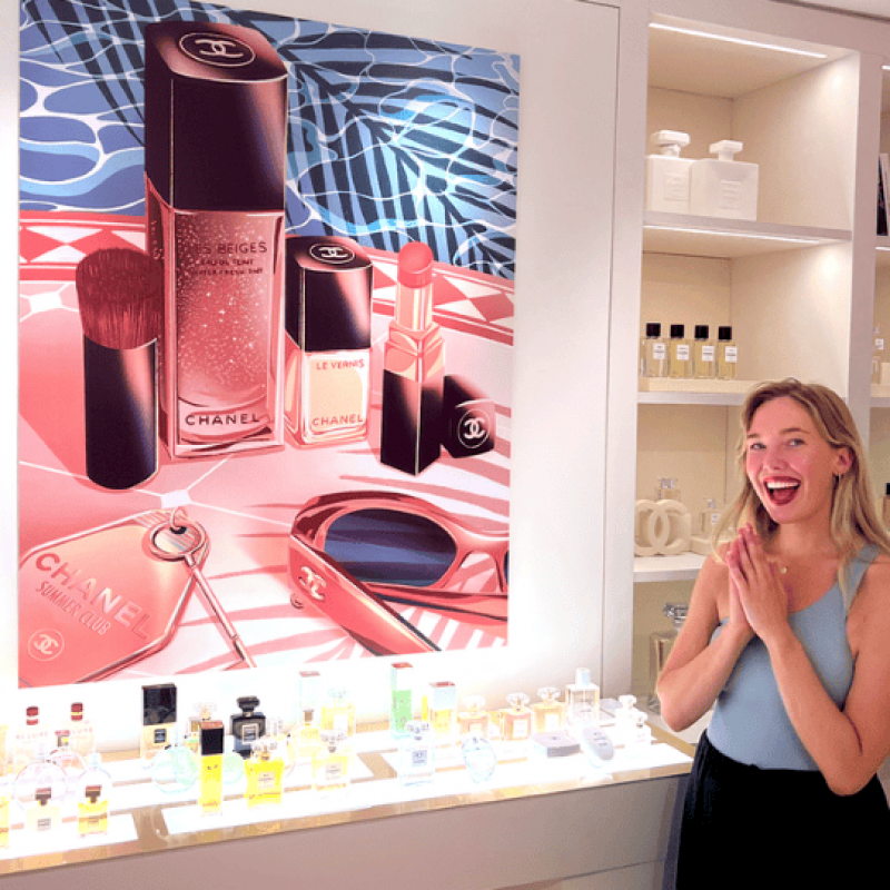 Izzy stands in a cosmetics store with prominently displayed Chanel products and her colourful designs with an excited expression on her face.