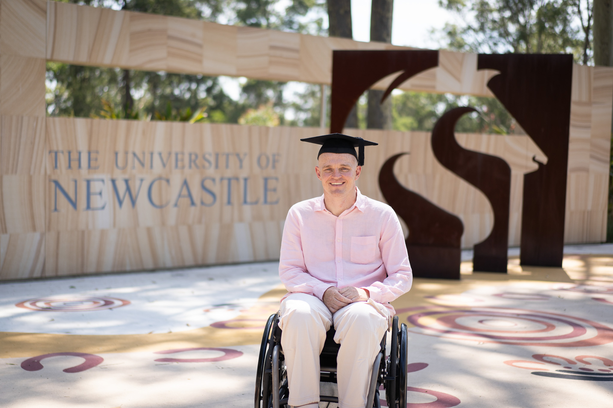 Alex McKinnon in a graduation cap in front of the University of Newcastle sign