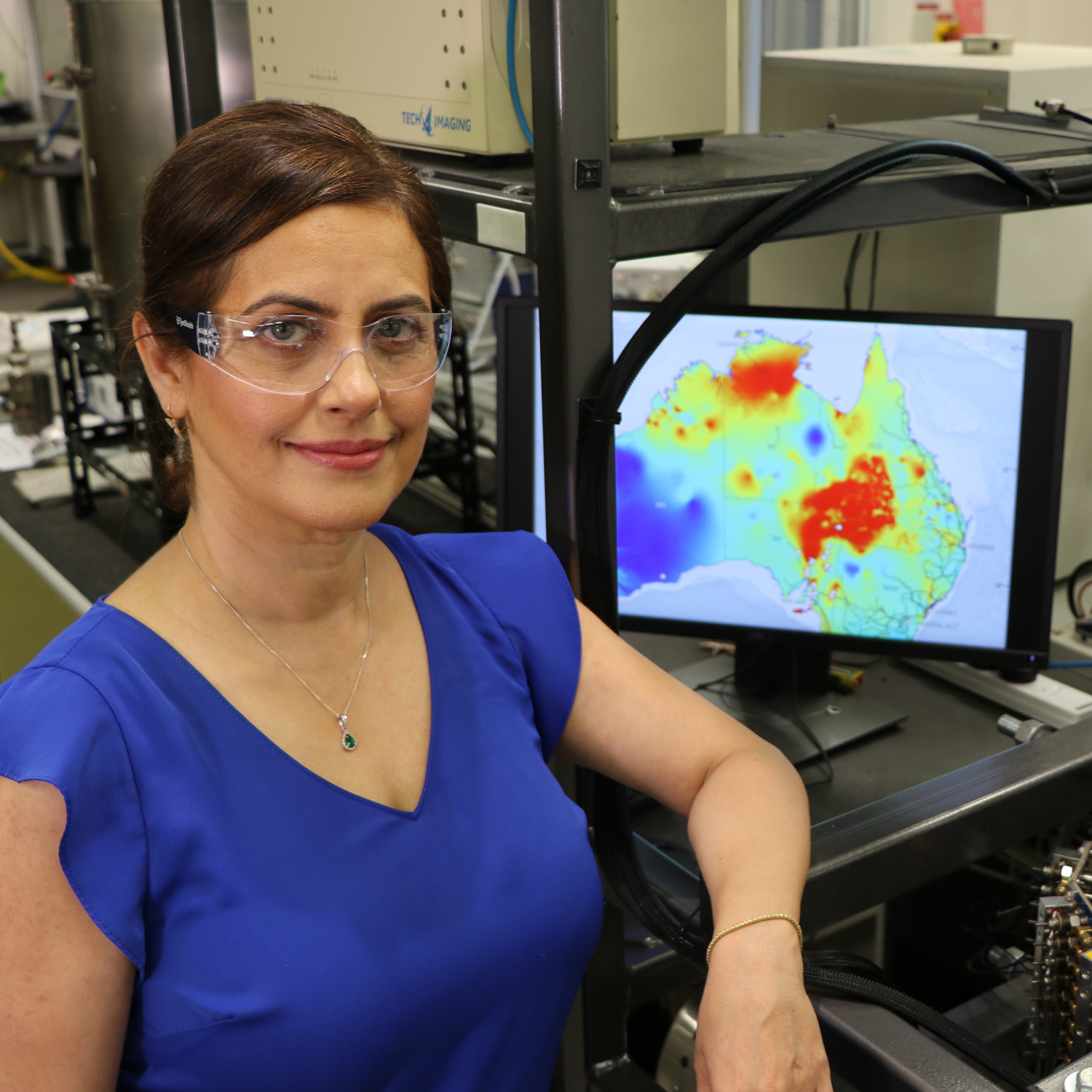 The researcher, Associate Professor Elham Doroodchi, is pictured smiling at the camera with safety goggles on in a lab environment. A diagram showing Australia heat spots is on a computer screen in the background