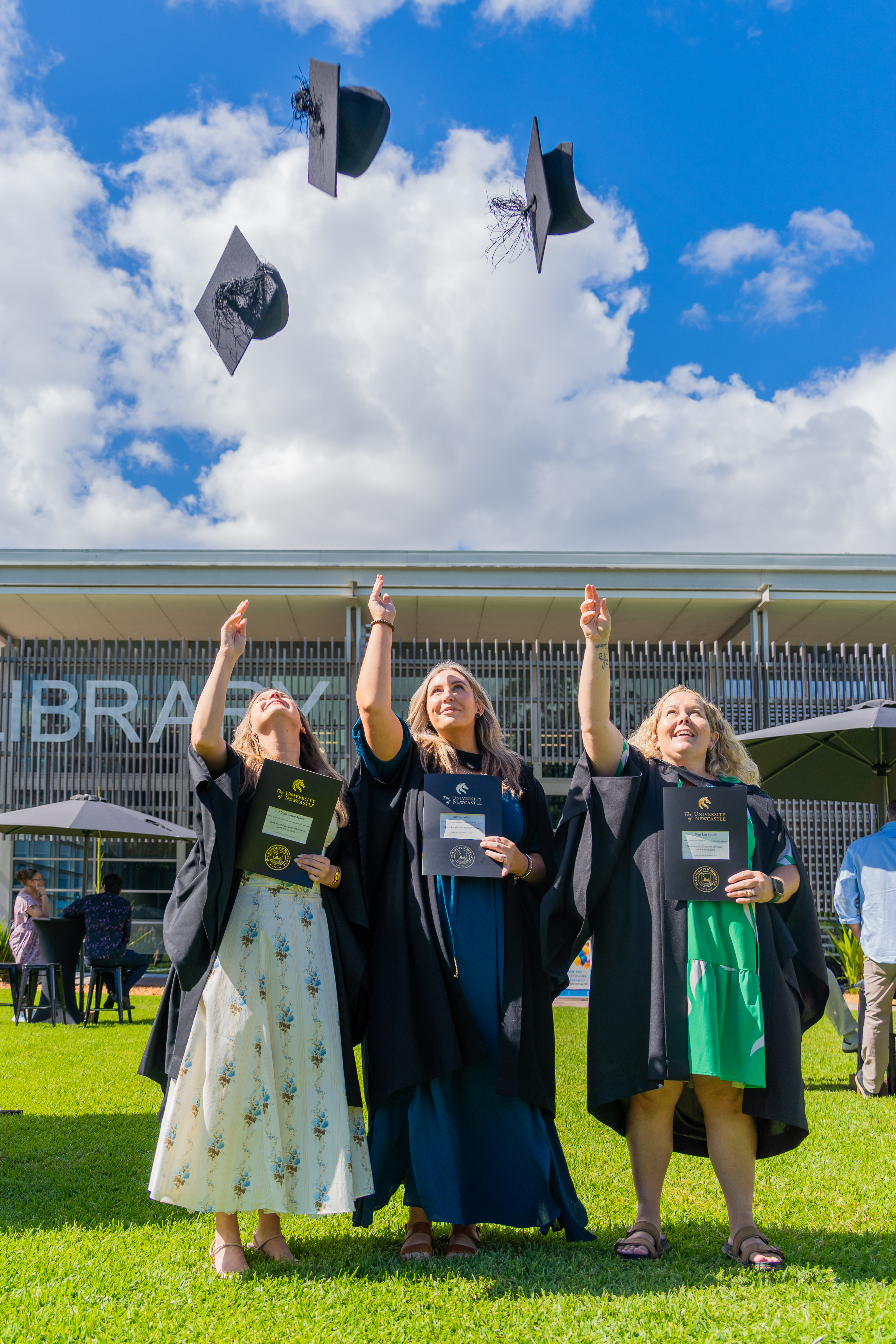 Three graduates in their gowns holding their certificates throw their graduation caps into the sky