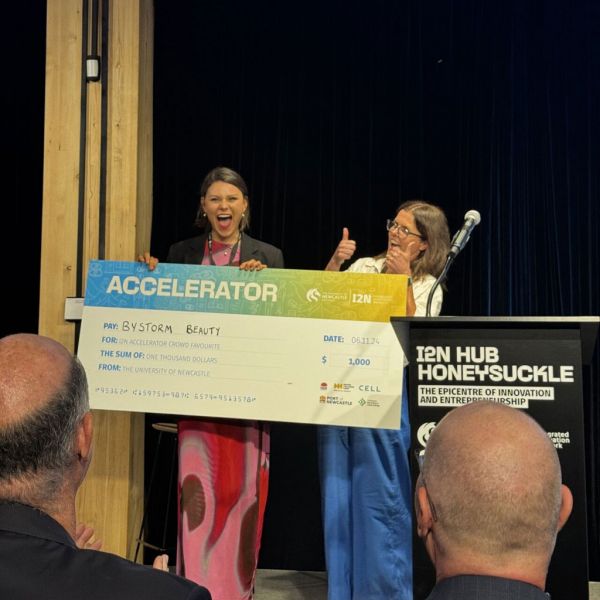 Storm stands by a podium alongside Siobhan Curran holding an oversized promotional check from the I2N Hub Honeysuckle Accelerator.