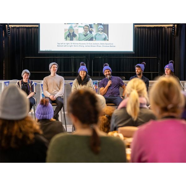 An image featuring six people sitting in a row on a stage all wearing Mark Hughes Foundation Beanies, looking out to a crowd. Six guests are captured in the foreground sitting on chairs listening. They are blurred for effect. From left to right. A person is looking down smiling. They have their hands in their lap and are wearing a grey beanie with a black tee and a striped shirt. A man is smiling looking out to the crowd. He’s wearing a grey beanie and a sand long sleeved knit. His hands are resting on his lap. A woman smiles directly to the audience. She has long, dark hair and is wearing a lilac beanie and a pale cardigan. A man is holding a microphone and smiling at the audience. He has a dark beard and is wearing a short-sleeve button up shirt and a lilac beanie. A man is looking out to the audience. He is wearing a dark collared shirt and a lilac beanie. A lady is looking out to the audience. You can only see her head as the audience are blocking the remainder of her body in this view. The foreground is showing 6 members of the audience listening to the panel speak. This part of the image is blurred.