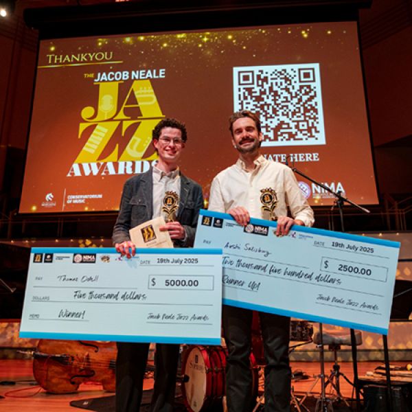 Two individuals stand on stage holding large checks for winning and runner-up positions at the Jacob Neale Jazz Awards, with a backdrop displaying event details and a QR code.. Celebrating the next generation of jazz musicians at the inaugural Jacob Neale Jazz Awards.