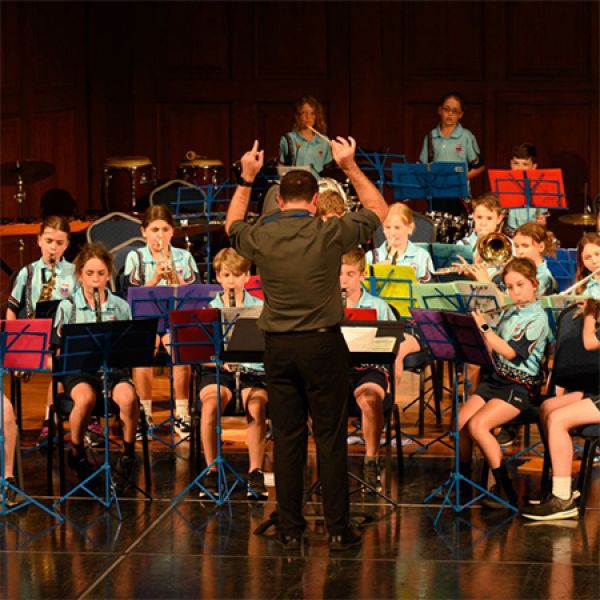 A conductor leads a uniformed band of seated musicians performing on stage with instruments and music stands.