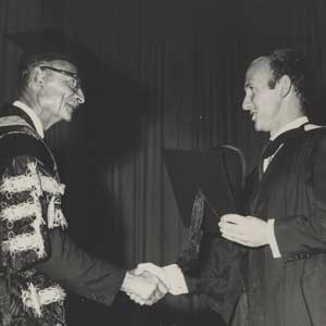 A Professor in a gown and cap shaking the hands of a graduate in a gown