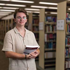 A student looks to camera while holding a textbook, with the library behind them