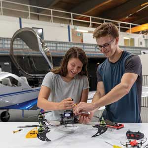 Teo engineering students standing in a warehouse with a small plane behind them, working on a drone using a tool, with a remote and other drone nearby