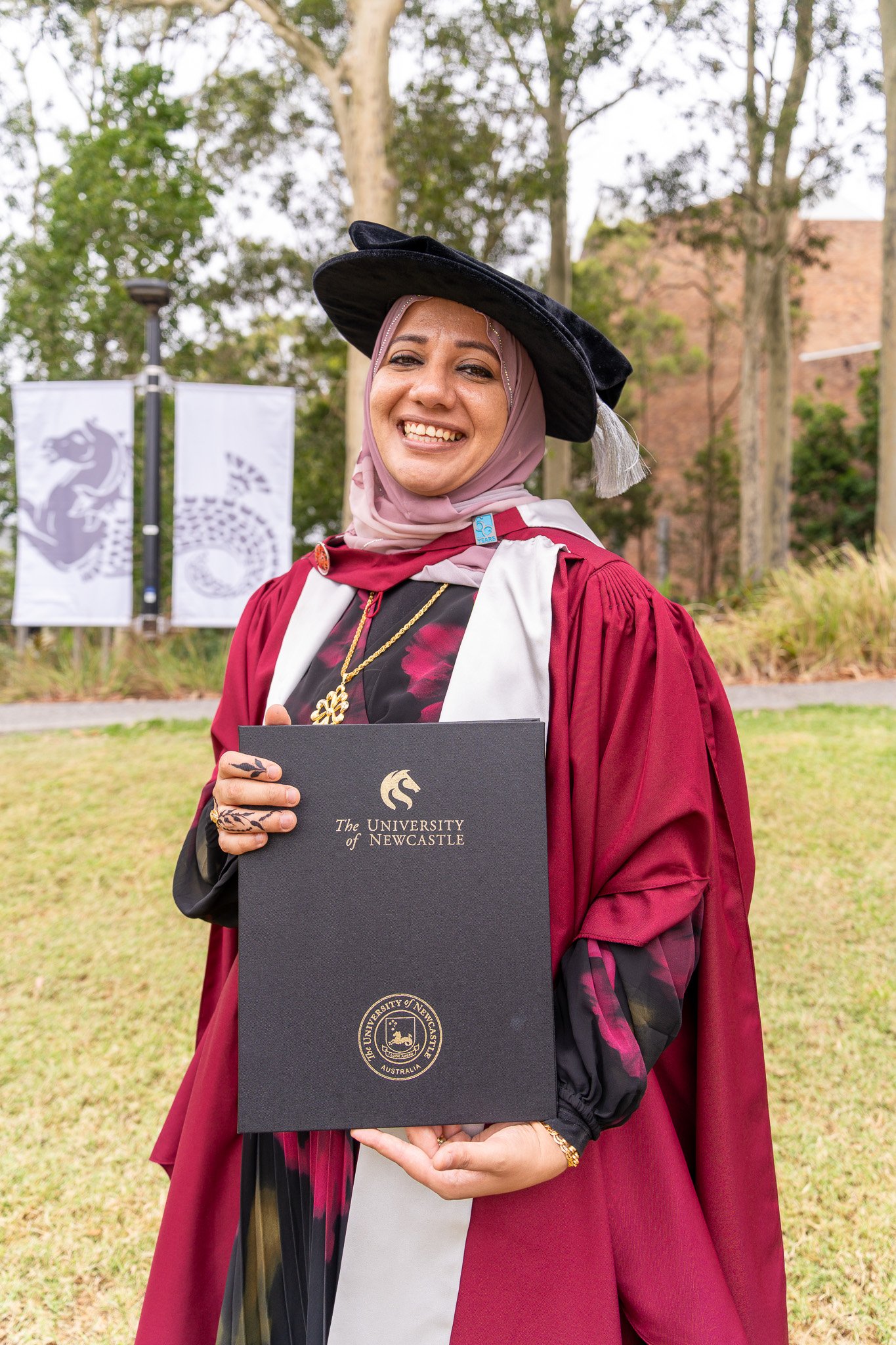 Dr Suzan Makhloof stands smiling with her testamur in hand and PhD graduation gown and hat on