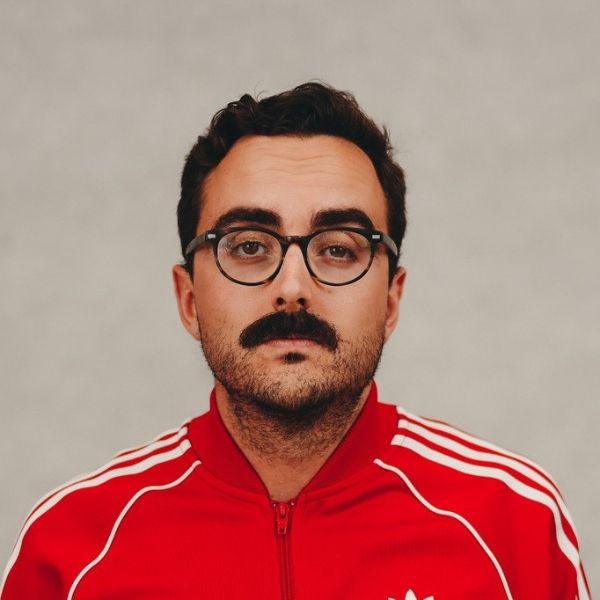 Studio portrait of a person with short dark hair, round glasses, and a thick moustache, wearing a red zip-up track jacket with white stripes against a plain light background.