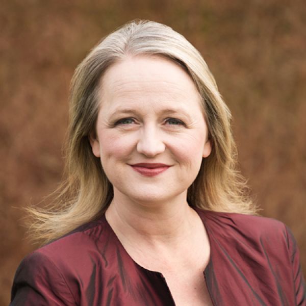 A close-up outdoor portrait shows a person with shoulder-length light hair wearing a maroon jacket, standing in front of a softly blurred brown background.